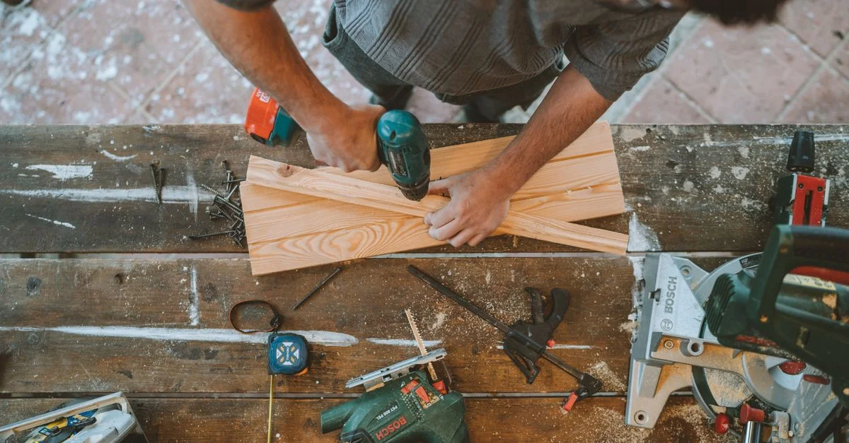 Artisan occupé à percer des planches de bois dans un atelier de menuiserie avec divers outils autour.
